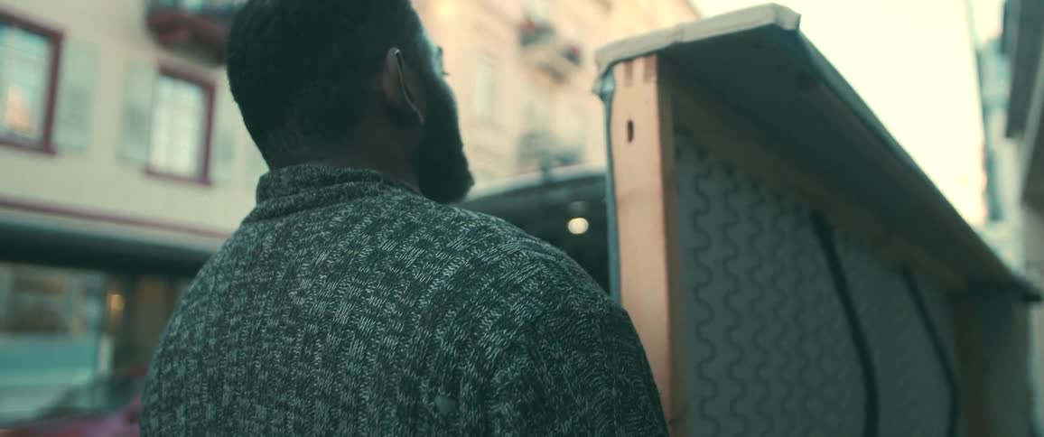 Over-the-shoulder slow-motion shot of a man carrying a piece of furniture down an urban street towards a moving van