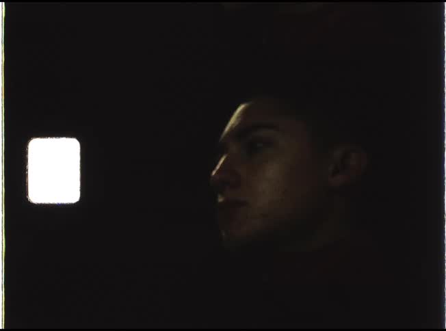 Vintage footage shows the profile of a pensive young man sitting in a dark room, faintly illuminated by a small square light