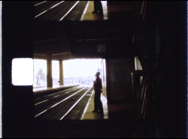 Archival film captures a lone figure waiting in silhouette by the tracks at a train station