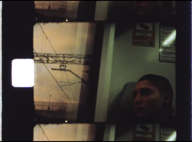 A thoughtful young person with a buzzcut is captured on Super 8 film while riding a train through an industrial area at dusk