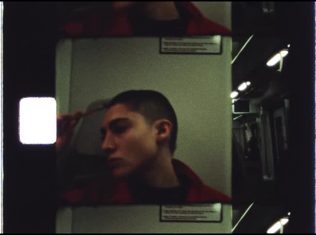 A vintage-style film captures a person with a shaved head meticulously applying eyebrow makeup while riding on a train