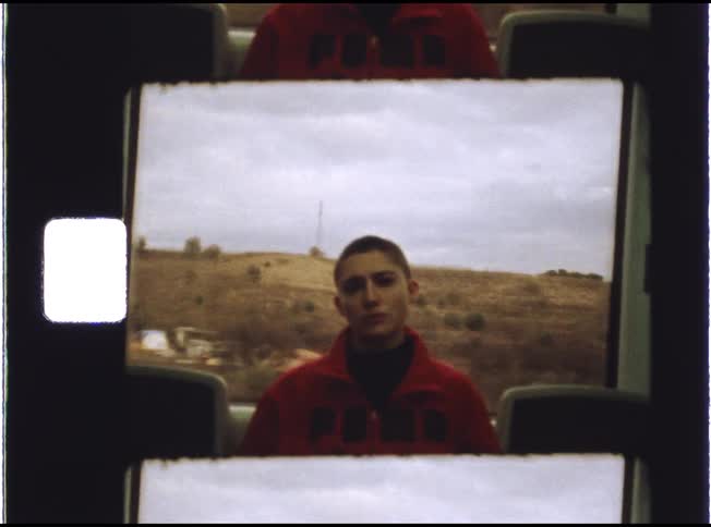 A vintage film scan captures a person with a buzzcut and a serious expression traveling on a bus through a rural landscape