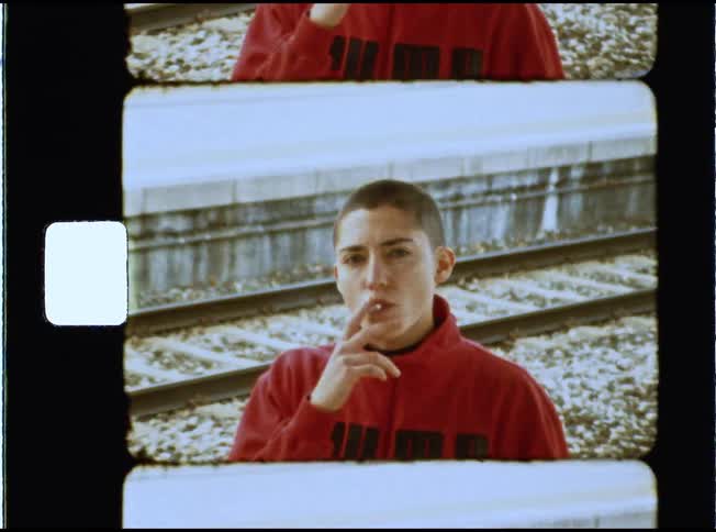 Vintage Super 8mm footage of a young person with a buzz cut lighting and smoking a cigarette at a train station while looking intently at the camera