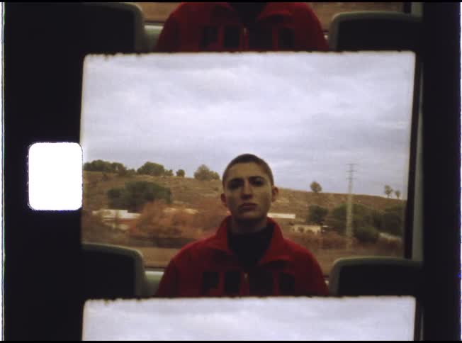 A pensive woman with a buzz cut stares into the camera as the countryside passes by her window on a moving vehicle, framed by a Super 8mm film strip