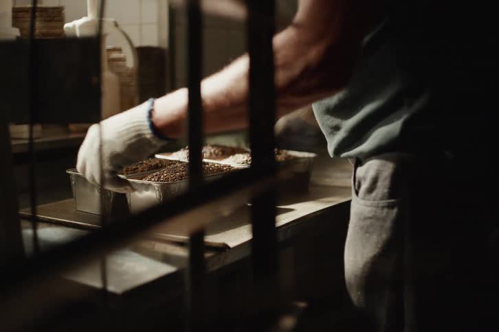 In a rustic bakery, a worker wearing gloves un-pans freshly baked loaves of seeded bread