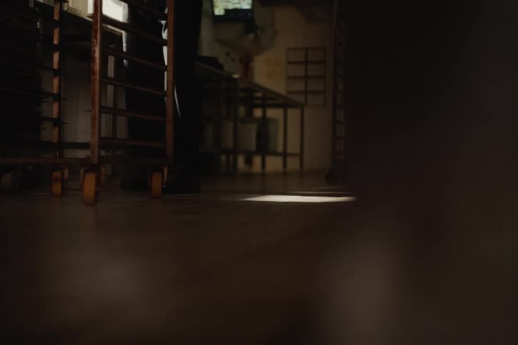 Low angle shot of a baker pushing a wheeled baking rack across the floor of a dimly lit kitchen
