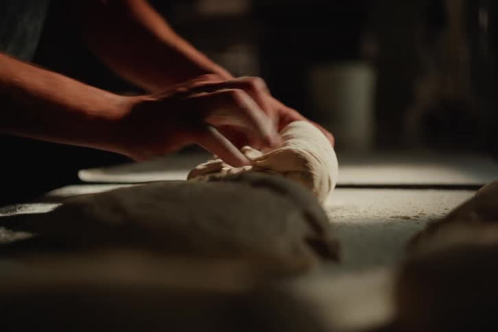 Close-up of a baker's hands skillfully kneading fresh dough on a floured countertop in a dimly lit kitchen