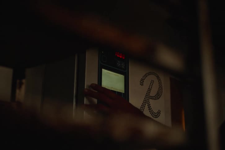 A person's hand sets the digital controls on an industrial oven in a dark bakery, preparing it for baking