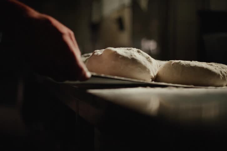 A baker in a dark kitchen places raw dough balls on a floured surface in slow motion