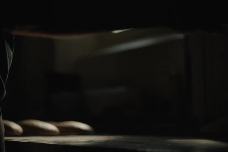 A baker's silhouette sprinkles flour across a wooden counter in a dimly lit room, with loaves of bread dough resting in the background
