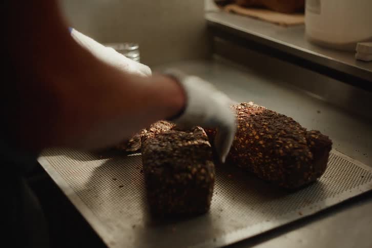 A baker carefully places freshly baked loaves of rustic, seeded bread onto a cooling tray