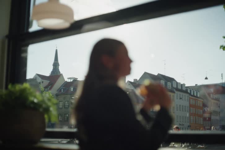 An out-of-focus woman sits by a window, enjoying the sunlit view of a historic European city