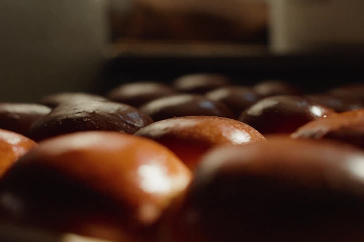 Warm sunlight illuminates rows of shiny, freshly baked brioche buns on a baking sheet