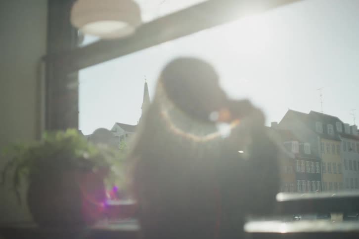 A sun-drenched view of a historic European city from a cafe window, with a woman's soft-focus silhouette in the foreground