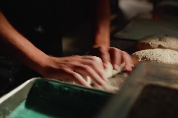 A baker skillfully kneads and shapes bread dough on a metal countertop in a dimly lit, rustic bakery