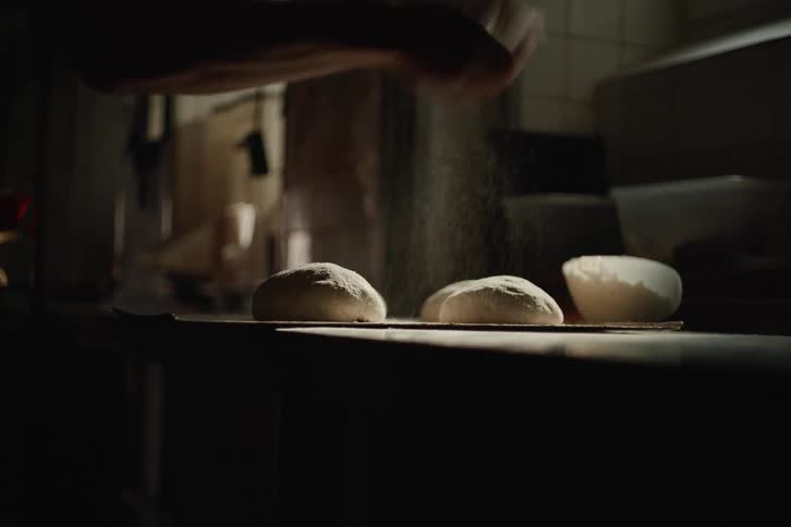 Slow motion shot of a baker's hands dusting fresh dough balls with flour in a dimly lit, atmospheric bakery