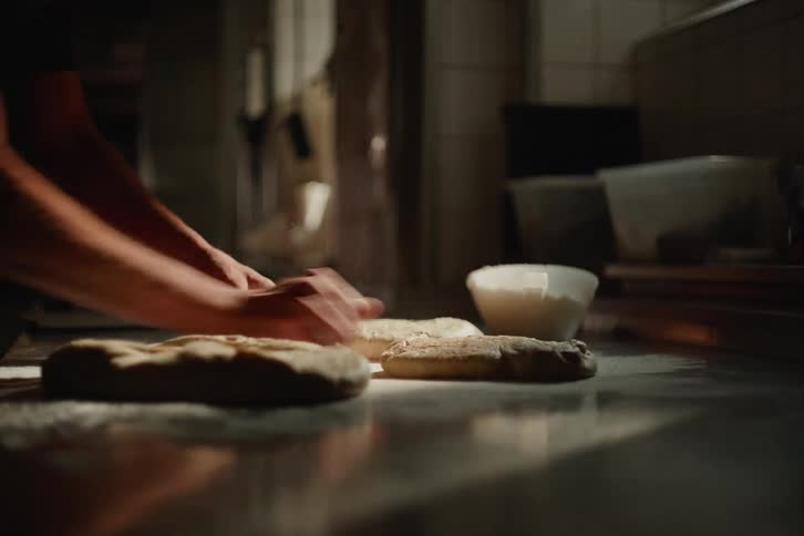 An artisan baker's hands expertly knead and fold fresh dough on a floured steel countertop in a dimly lit kitchen
