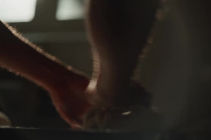 A close-up slow-motion shot of a baker's hands skillfully kneading dough on a floured surface in a dimly lit kitchen with soft backlighting