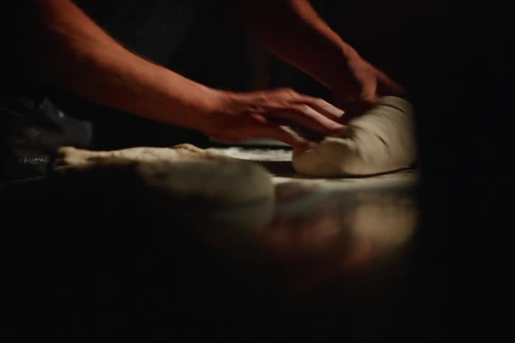 An artisan baker's hands skillfully knead, roll, and shape fresh bread dough on a floured steel countertop in a dark, atmospheric kitchen