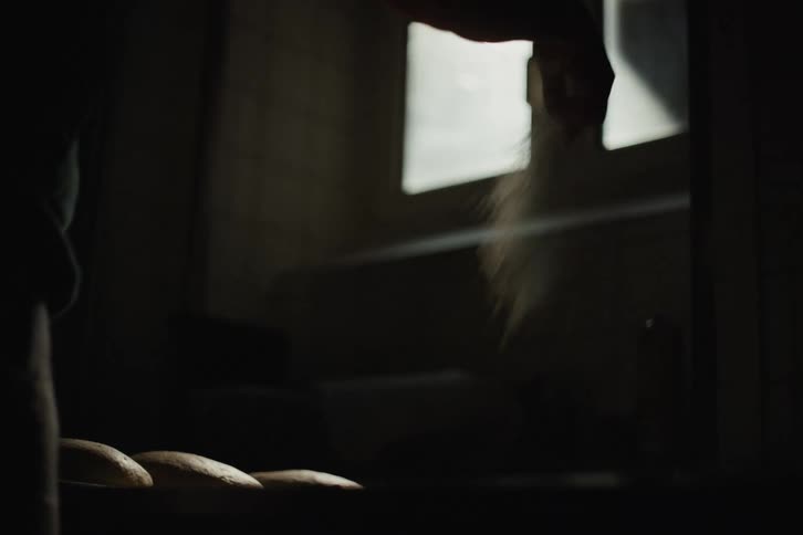 A baker sprinkles flour over fresh dough in a dark kitchen, beautifully captured in slow motion with dramatic backlighting
