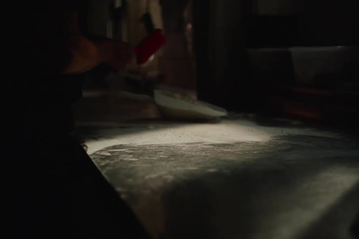 In a dimly lit kitchen, a worker meticulously sweeps flour off a stainless steel counter into a dustpan in slow motion