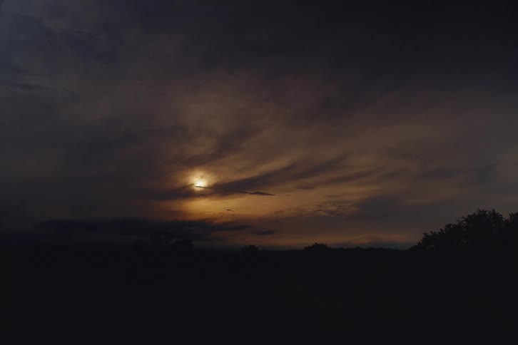 The sun glows behind dark, dramatic clouds during a moody sunset over a silhouetted landscape
