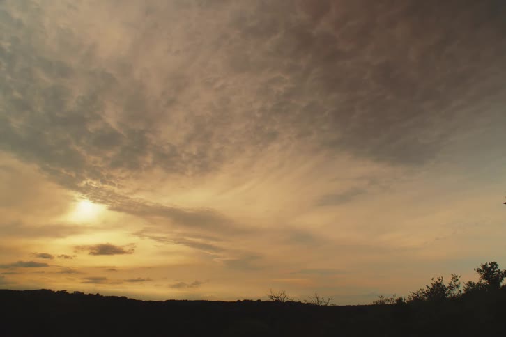A wide, static shot of a beautiful, warm sunset with scattered clouds filling the vast sky above a silhouetted horizon
