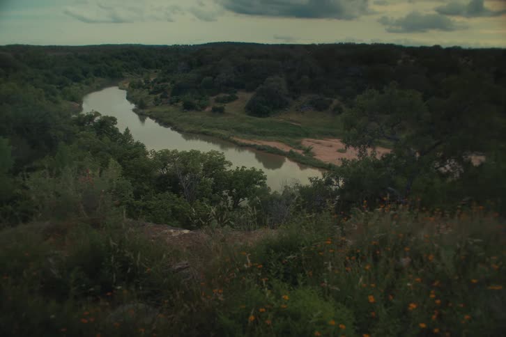 A wide, static shot captures a serene river winding through a lush green valley under a moody, overcast sky, with wildflowers in the foreground
