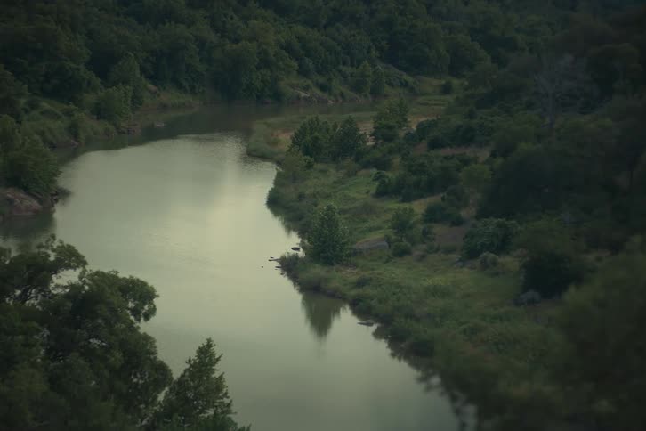A high-angle, static shot of a calm river winding through a dense, lush green forest at twilight