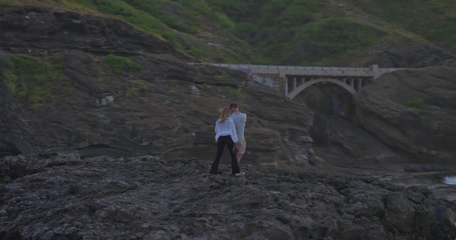 A romantic couple embraces on a rocky coastal cliff overlooking the ocean as the camera slowly orbits them at sunset