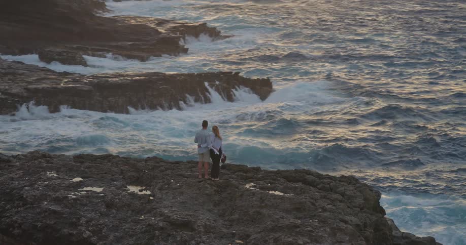 A loving couple stands on a rocky cliff, embracing as they watch the waves of the vast ocean during a beautiful golden sunset