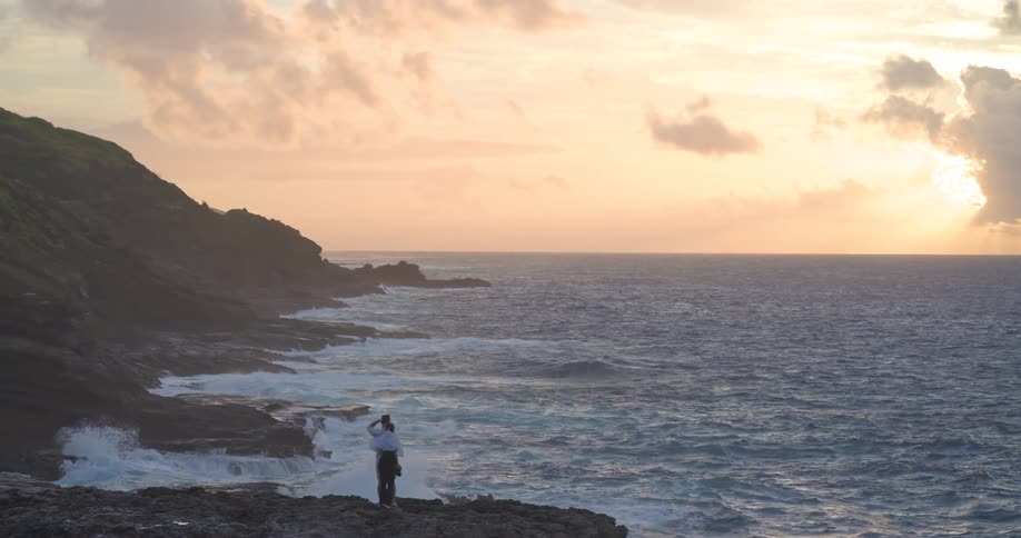 A romantic couple stands on a dramatic, rocky coastline, embracing as they watch the beautiful sunrise over the turbulent ocean