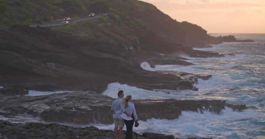 An aerial view reveals a romantic couple embracing on a dramatic rocky coastline, watching powerful waves crash below as the sun sets