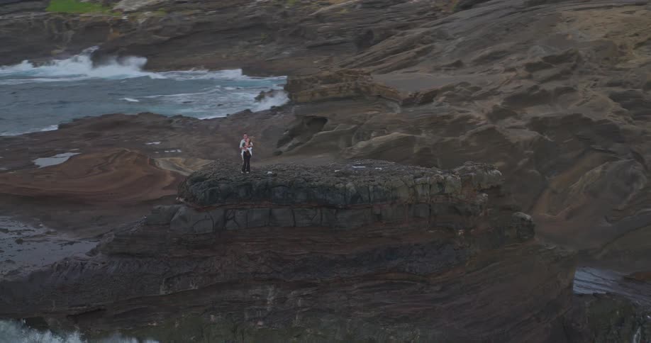 An aerial view shows a couple embracing on a dramatic rocky cliff overlooking the powerful, crashing ocean waves