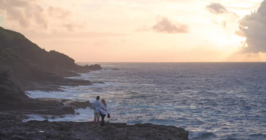 A romantic couple stands on a rocky cliff overlooking the ocean, embracing as they watch the beautiful sunset together