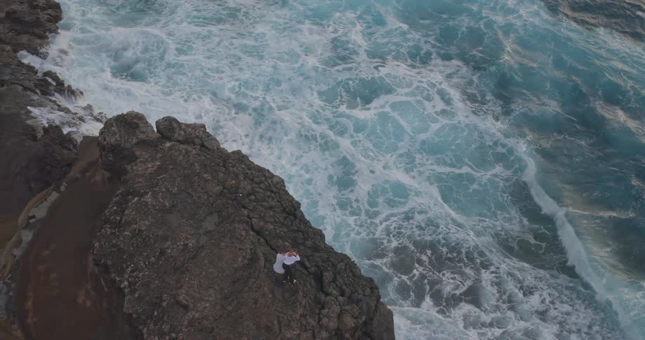 A dramatic aerial shot follows a couple standing on a rugged cliff overlooking the powerful crashing waves of the ocean