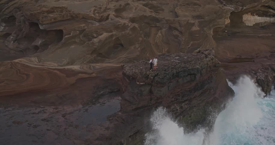 Epic aerial view pulls back to reveal a couple standing on a dramatic rocky coastline as powerful waves crash below