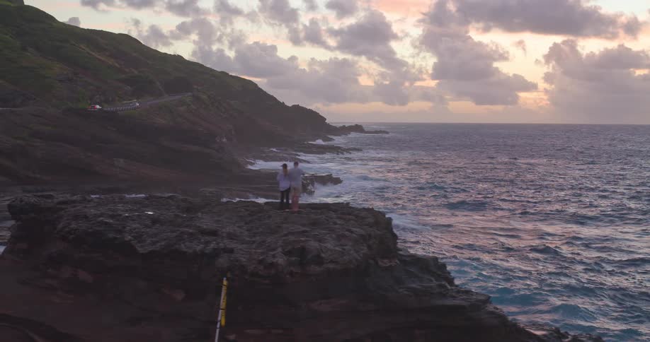 A drone pulls back from a beautiful sunrise over a rugged coastline to reveal a couple standing on a rocky cliff overlooking the vast ocean