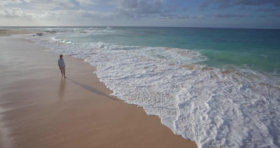 A man walks along a beautiful sandy shoreline as large turquoise ocean waves crash in slow motion