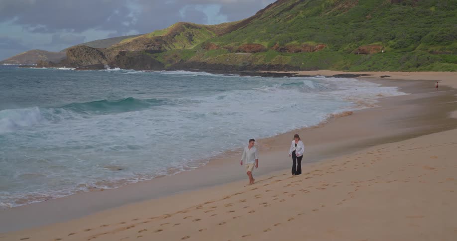 A couple enjoys a romantic walk along the shoreline of a beautiful sandy beach with green mountains in the background