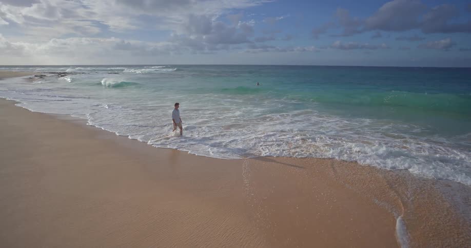 A man enjoys a peaceful walk in slow motion along a vast sandy beach as the turquoise ocean waves roll onto the shore