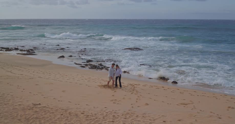 An aerial view follows a romantic couple as the man gives the woman a piggyback ride and they walk along a secluded sandy beach