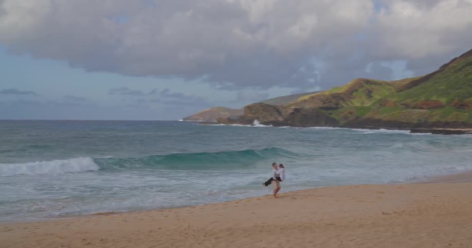 A man gives a woman a loving piggyback ride as they stroll along a scenic sandy beach with ocean waves and mountains in the background