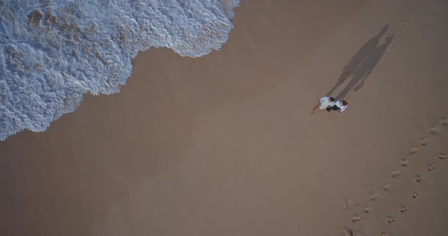 An aerial top-down slow-motion view of a romantic couple holding hands and walking along a sandy beach as the ocean waves gently roll ashore