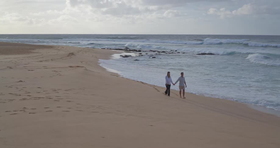 An aerial view of a couple holding hands and walking along a beautiful sandy beach as the ocean waves gently roll ashore