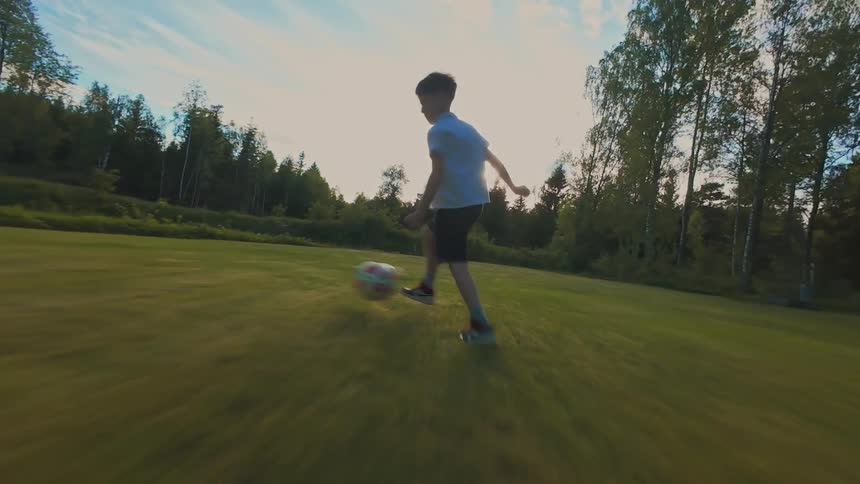 A young boy energetically kicks a soccer ball across a vibrant green lawn as the sun sets, captured in a dynamic, low-angle tracking shot