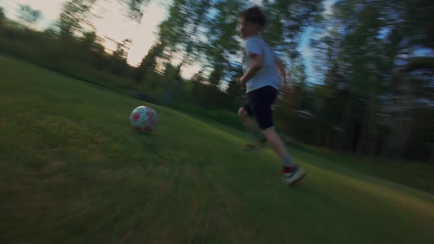 Energetic slow-motion follow shot of a young boy playing soccer in a lush green backyard at sunset