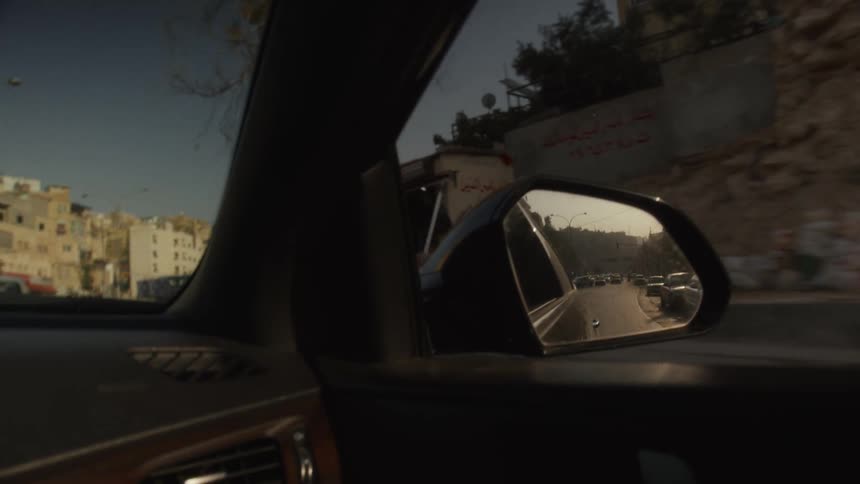 The road behind is reflected in the side mirror of a car driving through a sun-drenched Middle Eastern city
