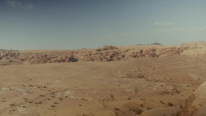 A wide, epic shot shows a small group of people trekking across a vast desert valley towards distant rocky mountains