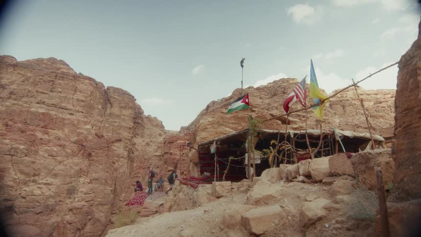 A small, rustic shelter decorated with international flags offers a resting spot for tourists in a Jordanian desert canyon